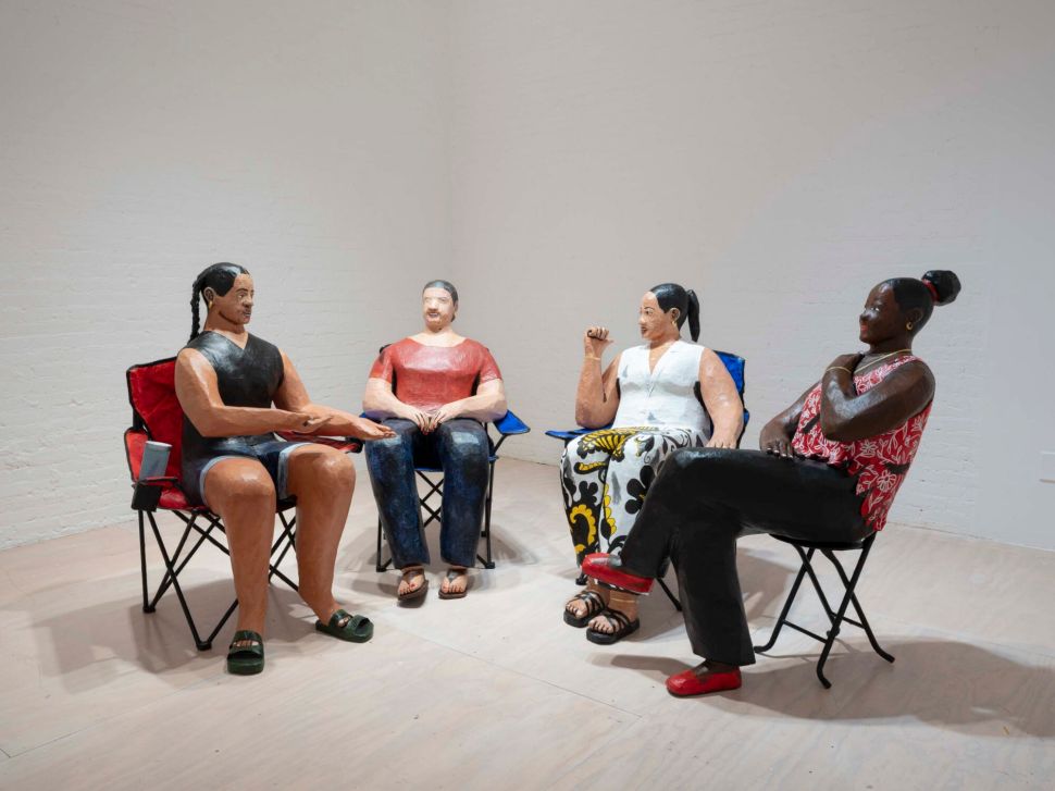 A group of four life-size painted sculptures of seated figures face each other in folding chairs arranged in a circle in a white gallery space.
