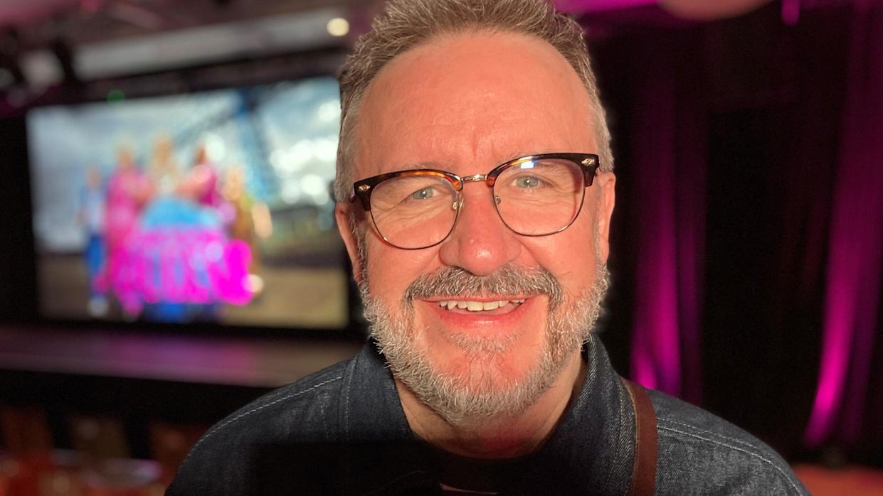 Mark Benton is smiling broadly and sitting down against a blurred but colourful poster of the show. He has short hair and a grey beard and is wearing glasses.