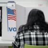 A person with their back to the camera marks their ballot in a voting booth bearing a U.S. flag and the word "Vote."
