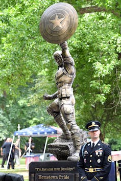 A bronze statue of a muscular superhero who's hoisting a shield with a star on it into the air.