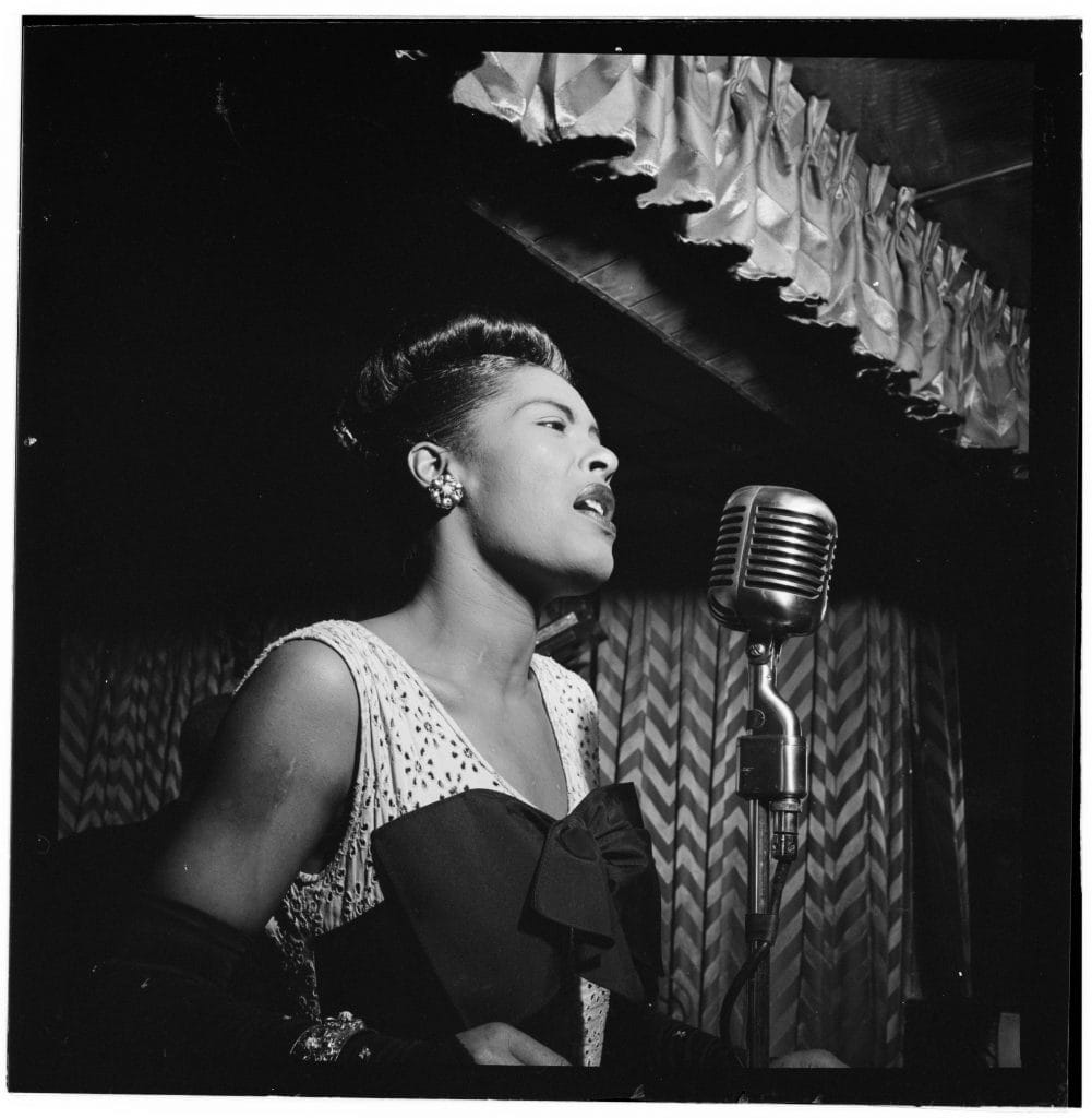 Black-and-white portrait captures Billie Holiday singing passionately into vintage microphone beneath softly draped nightclub curtains.