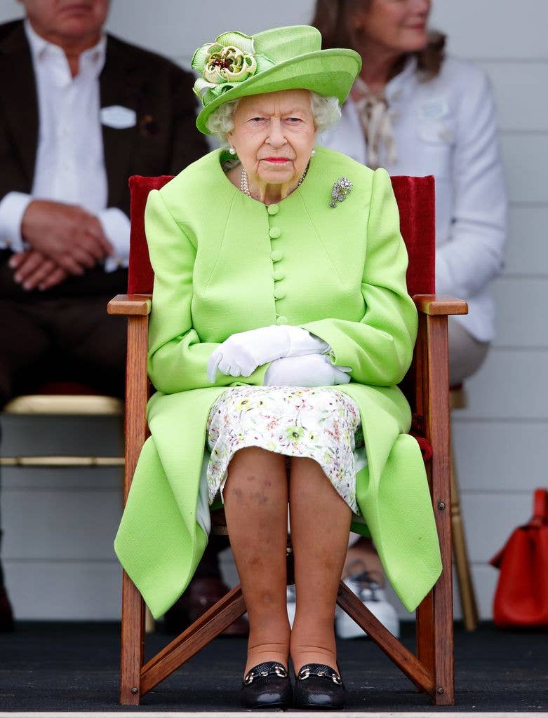 Queen Elizabeth II with a stern expression sitting with her arms crossed in a green coat