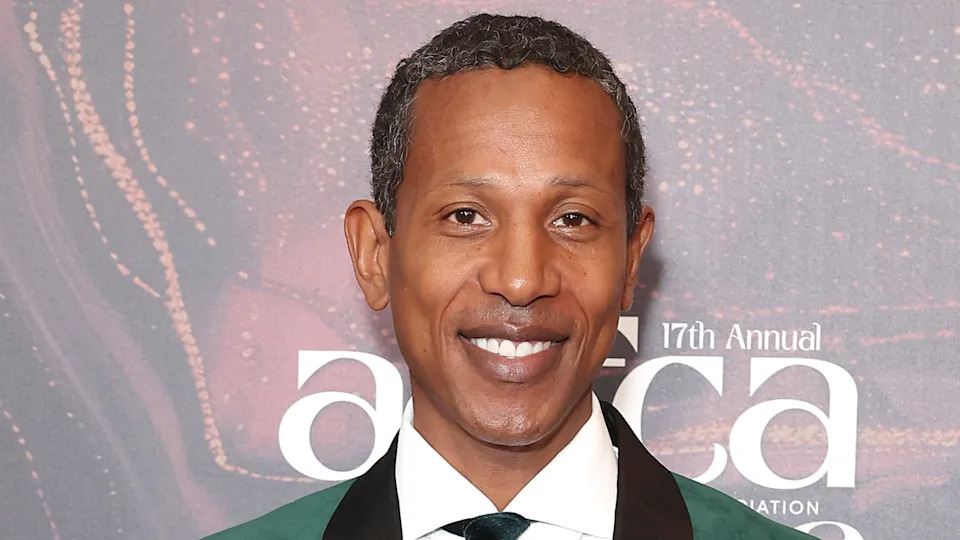 Shyne smiling at an event, wearing a green jacket with a black lapel and a white shirt, against a backdrop with text.