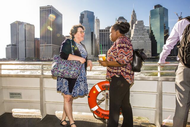 Two women stand on a ferry talking to each other with the Manhattan skyline in the background