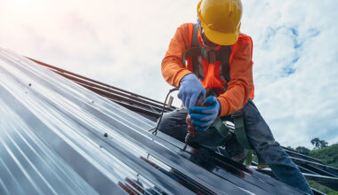 Roofer worker in special protective work wear and gloves,using air or pneumatic nail gun and installing asphalt or bitumen shingle on top of the Metal Sheet roof.