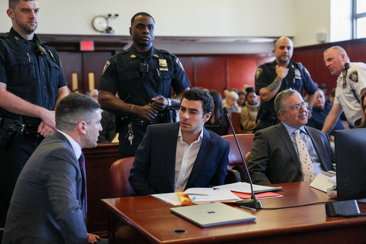A man sits at a table with his lawyers as police officers stand over him.