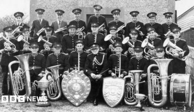 A black and white photo of Indian Queens Band assembled in front of their bandroom with several trophies and their instruments