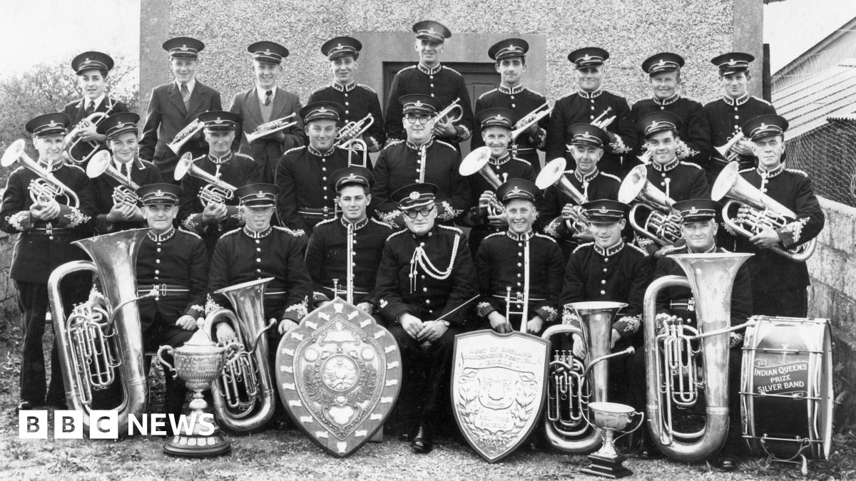 A black and white photo of Indian Queens Band assembled in front of their bandroom with several trophies and their instruments