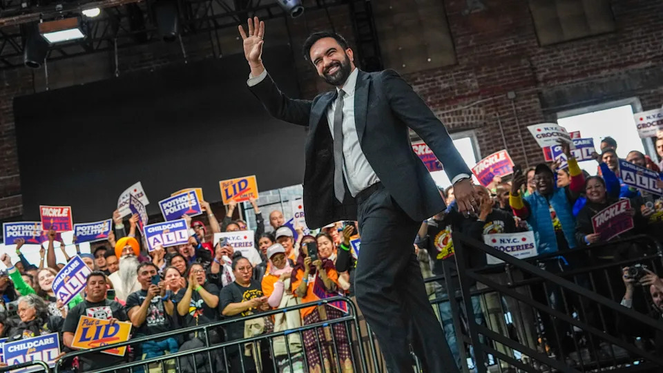 Zohran Mamdani in a suit waves to a cheering crowd holding signs in a large indoor venue.