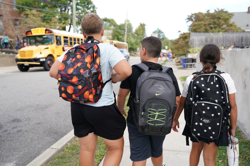 Three elementary school students wearing backpacks walk away from a school bus on their first day of classes.