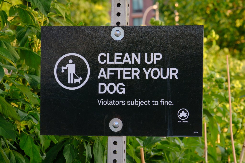 A black sign in a dog park reads "CLEAN UP AFTER YOUR DOG. Violators subject to fine."