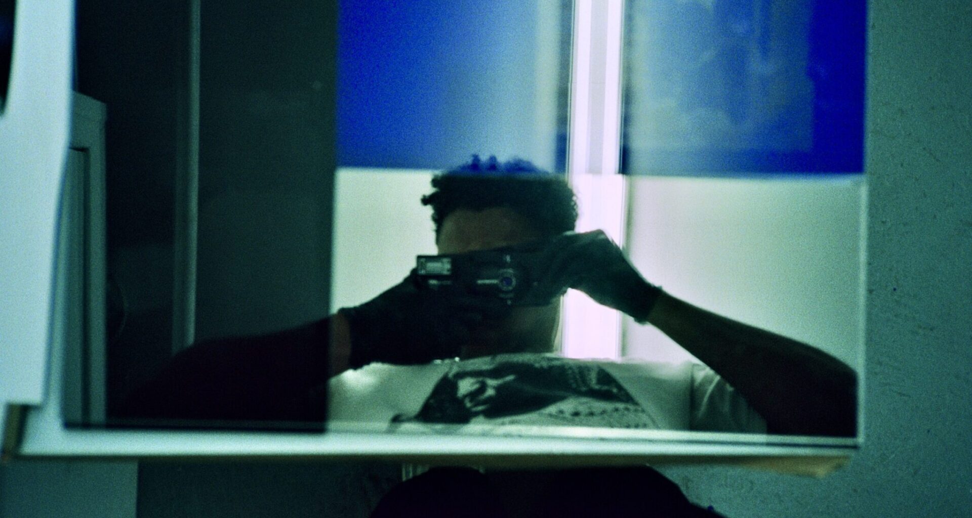 Artist takes a self-portrait in a mirror in a room with blue lighting, face obscured by the camera