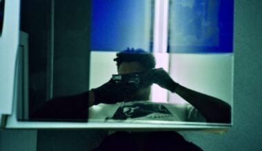 Artist takes a self-portrait in a mirror in a room with blue lighting, face obscured by the camera