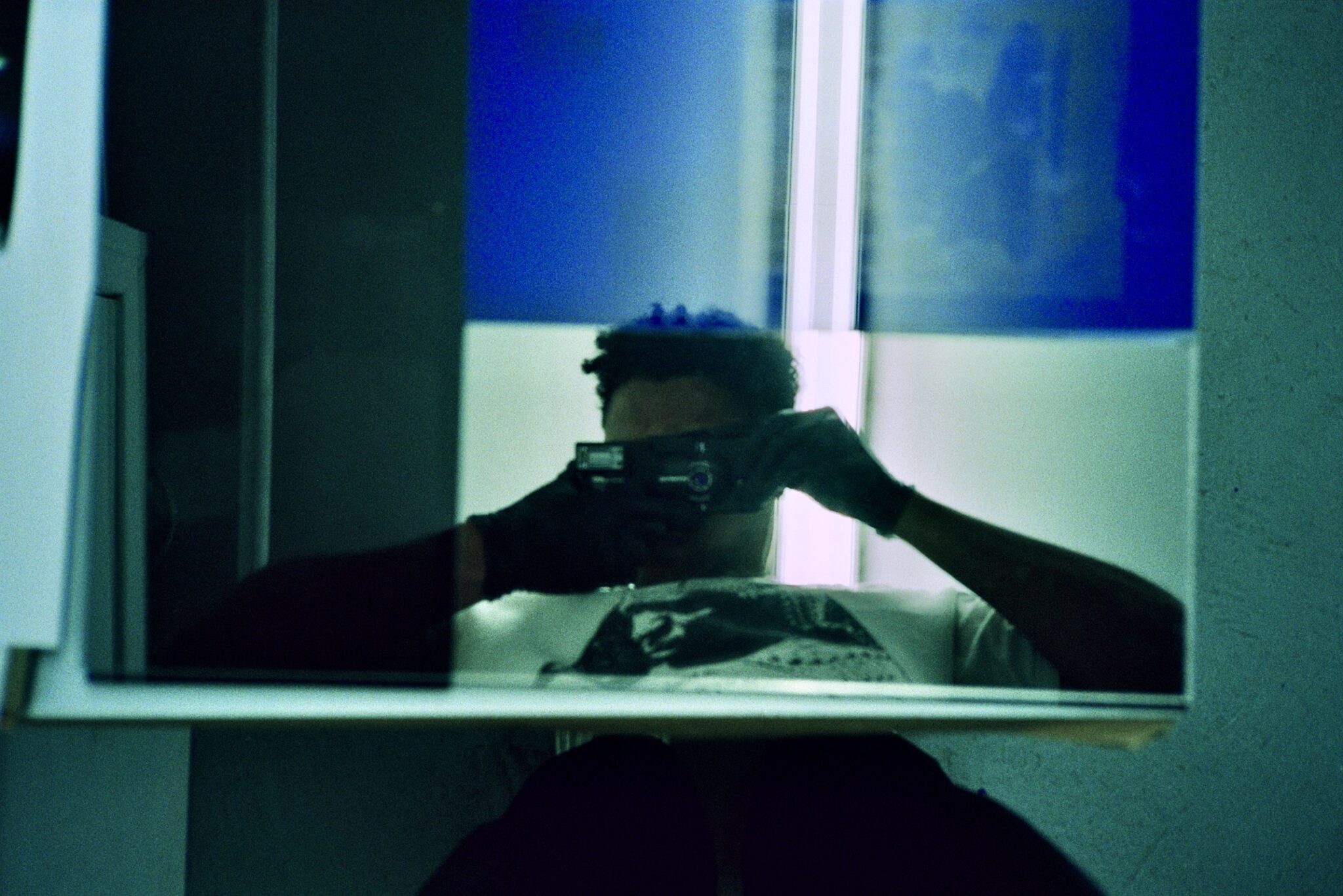 Artist takes a self-portrait in a mirror in a room with blue lighting, face obscured by the camera