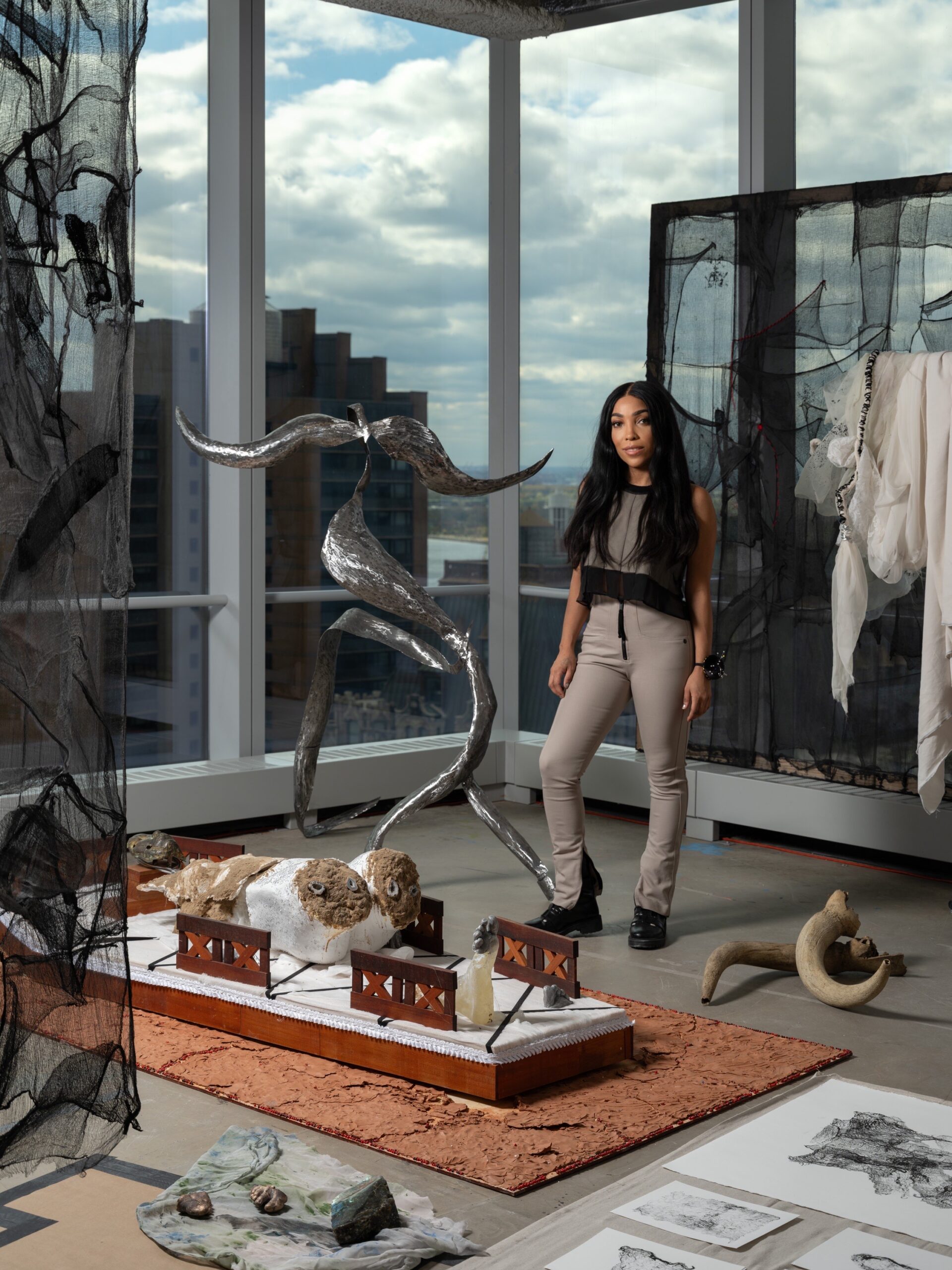 Dana-Marie Bullock stands in front of sculptural works in an airy contemporary space on a high floor of a tower with floor-to-ceiling windows