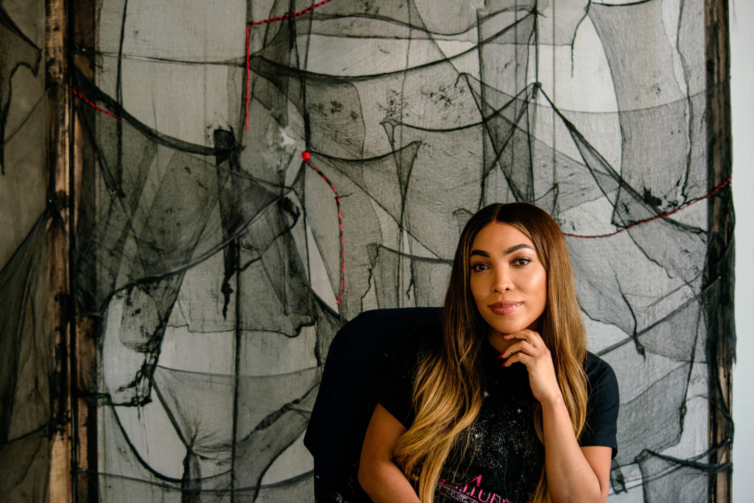 Alumna Dana-Marie Bullock seated with chin in hand in front of work comprised of interwoven sheer black pieces of fabric