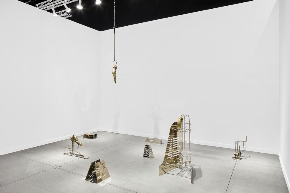 A sparse white booth presents metallic sculptural fragments arranged across the floor and suspended from above, casting delicate shadows on the walls.