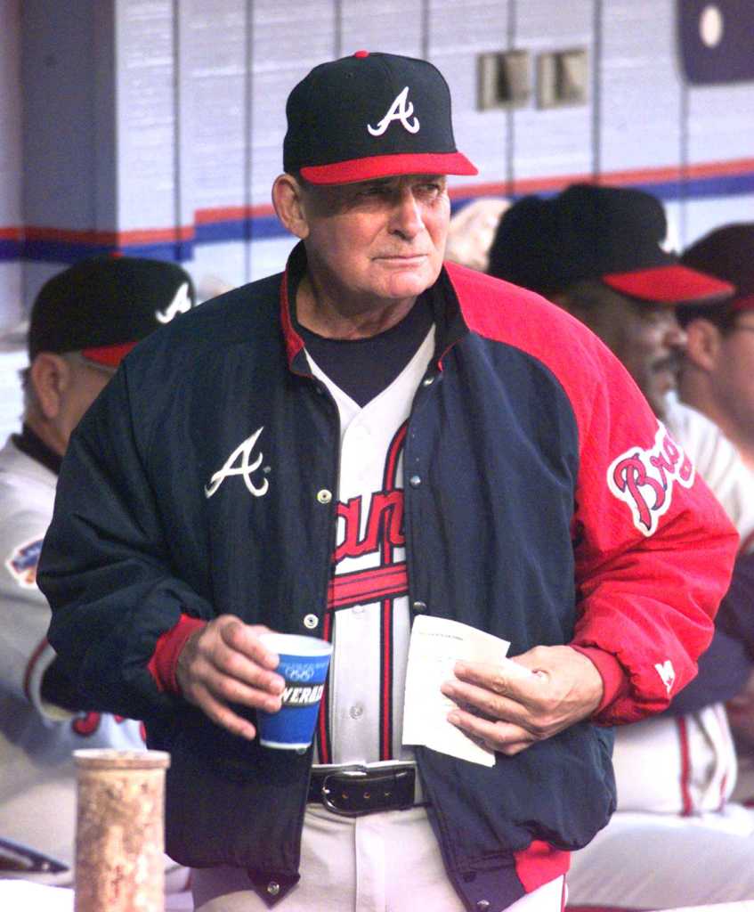 Bobby Cox in a Braves uniform, holding a cup and papers in the dugout.