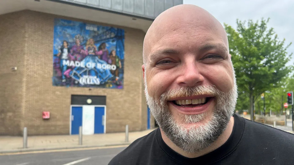Stephen Irving smiling for the camera with the mural in the background behind him. He is bold and has a short salt and pepper beard. He has blue eyes and is wearing a black t-shirt.