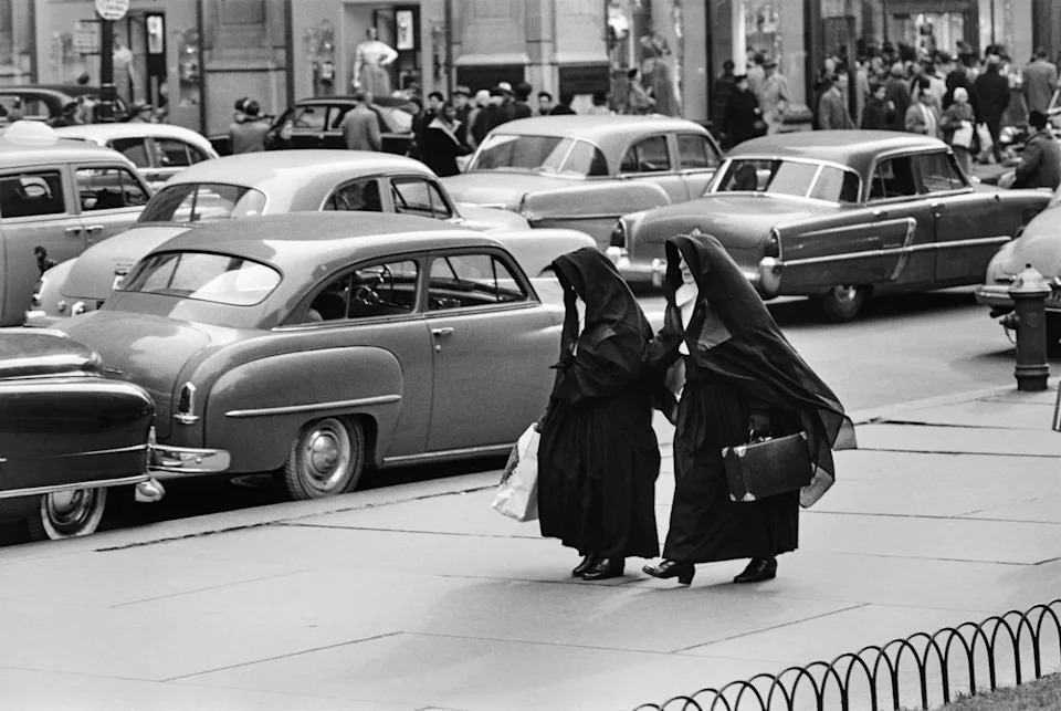 Two nuns in full black habits lean in towards each other in close conversation on a Midtown pavement, surrounded by the gleaming chrome and curves of 1950s American cars.
