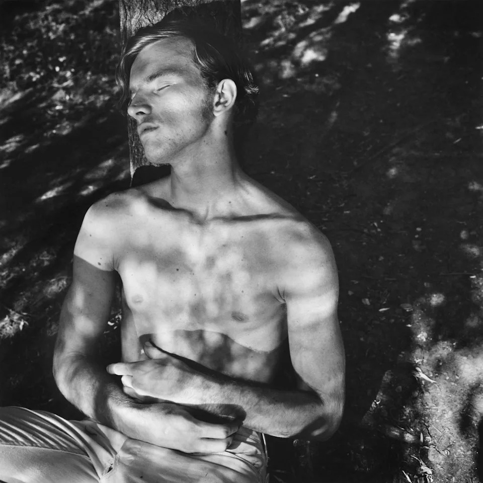 Black and white photo of a shirtless person sitting against a tree with shadows cast on their body and ground