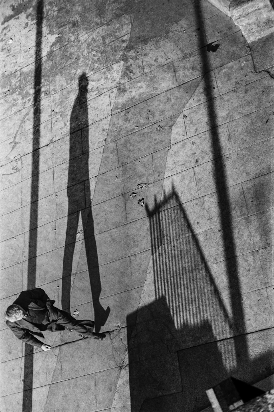 Shot from high above, a lone pedestrian in a coat is dwarfed by his own enormously elongated shadow and the falling shadow of a Manhattan skyscraper beside him.