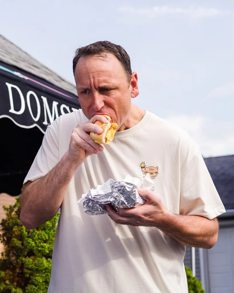 Joey Chestnut takes a bite out of a bacon, egg, and cheese bagel sandwich while holding another wrapped in foil. GOAT USA