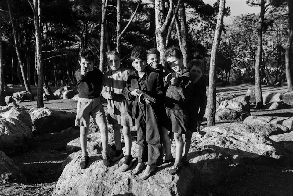 Five boys crowd together on a flat rock in a pine forest, pointing toy guns directly at the camera with grins that don't quite mask the aggression of the pose.