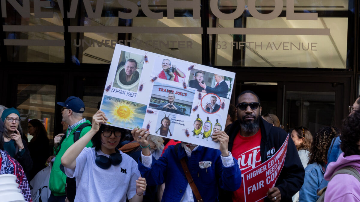 Alt Text: Protesters holding various signs stand outside a building with the words, University Center, 63 Fifth Avenue. Two students hold a humorous sign mocking Joel Towers, they stand next to a man in a red United University Professors' shirt. 