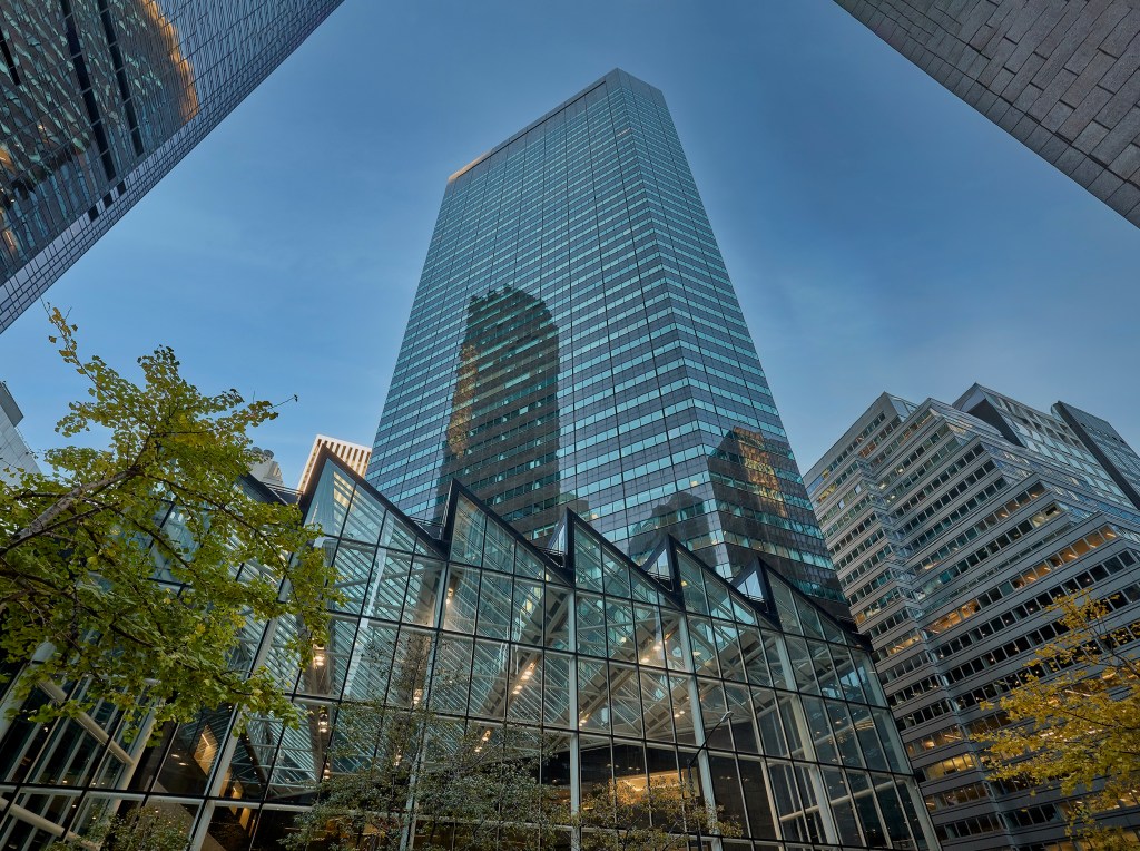 The 590 Madison Avenue skyscraper with a zig-zag glass atrium at its base, surrounded by other office buildings and trees.