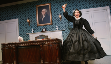A woman in a black period dress stands by a large wooden desk, holding up a teacup, with a portrait of George Washington hanging on the wall behind her.
