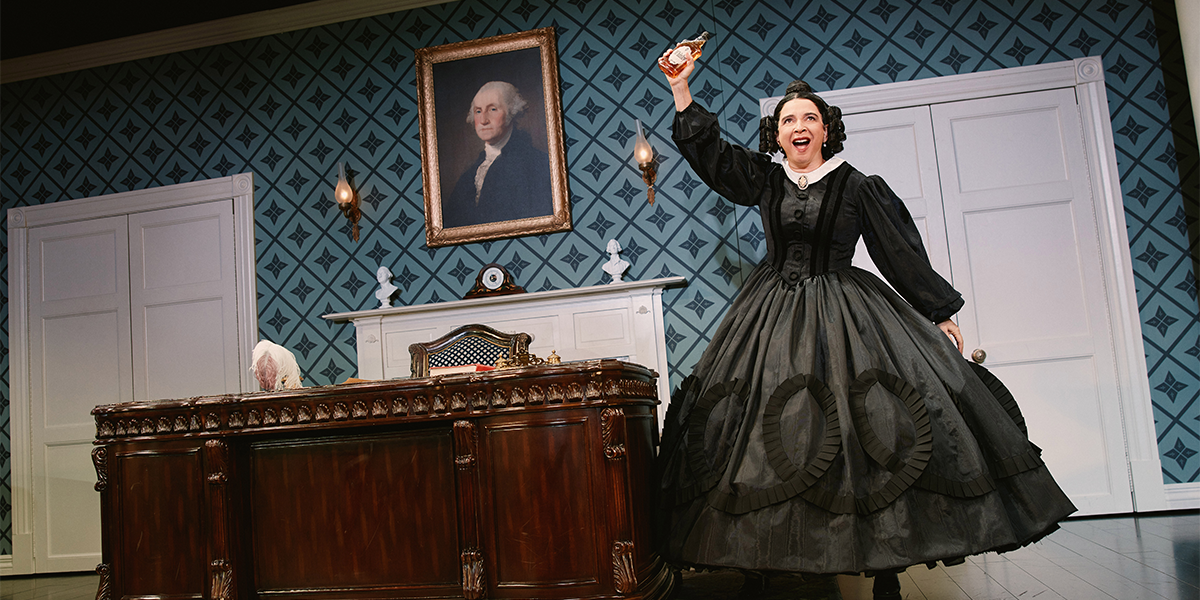A woman in a black period dress stands by a large wooden desk, holding up a teacup, with a portrait of George Washington hanging on the wall behind her.