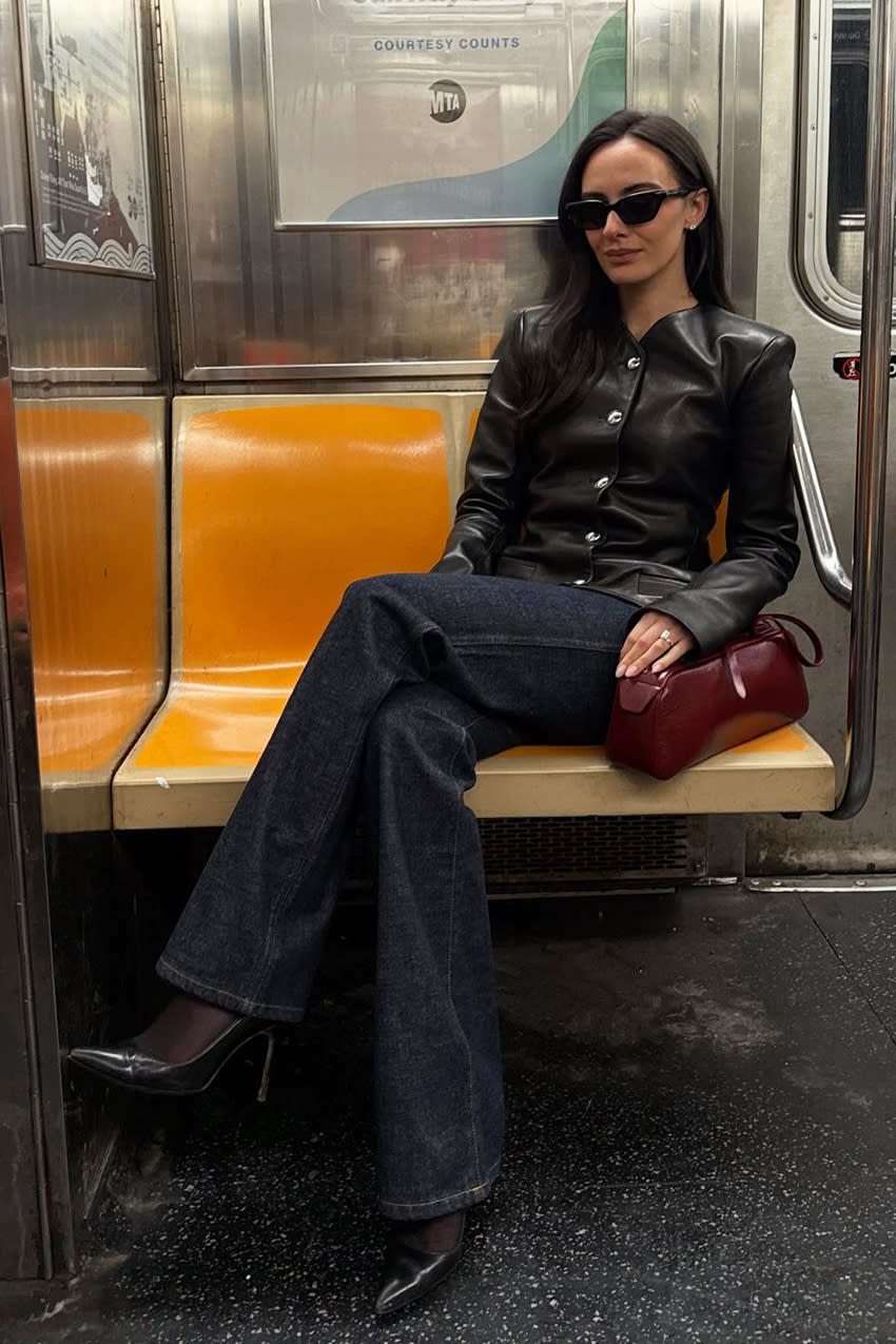 a woman on the subway wearing a black leather button-down jacket, dark-wash jeans, and black pointed-toe pumps