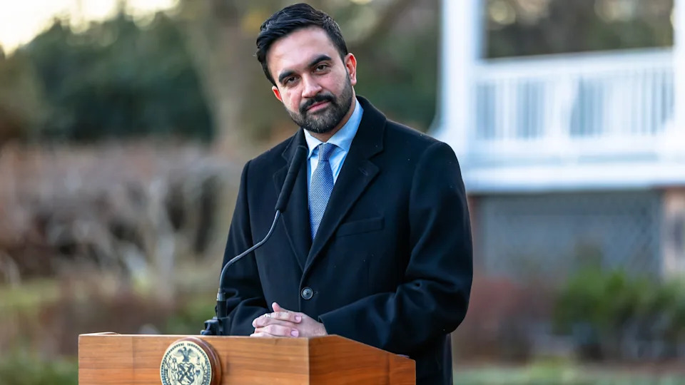 Zohran Mamdani in a dark coat and blue tie stands at a podium outdoors, with blurred greenery and a building in the background.