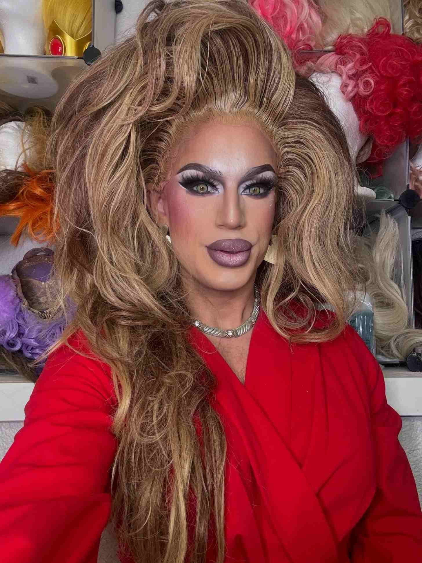 A drag queen takes a selfie, surrounded by wigs