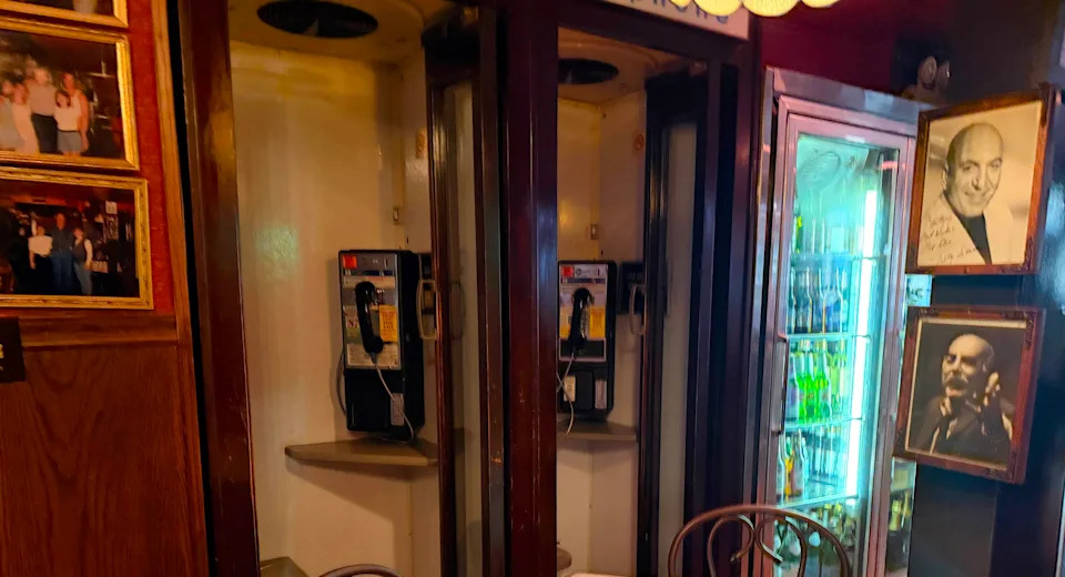 Two wooden phone booths with pay phones stand next to a built-in cooler filled with multi-colored beer bottles, both surrounded by walls adorned with photographs of families and celebrities.