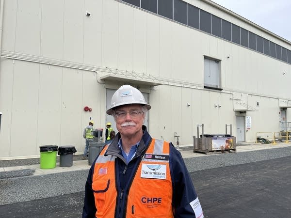 A man with a high visibility vest and helmet stands in front of an industrial building.