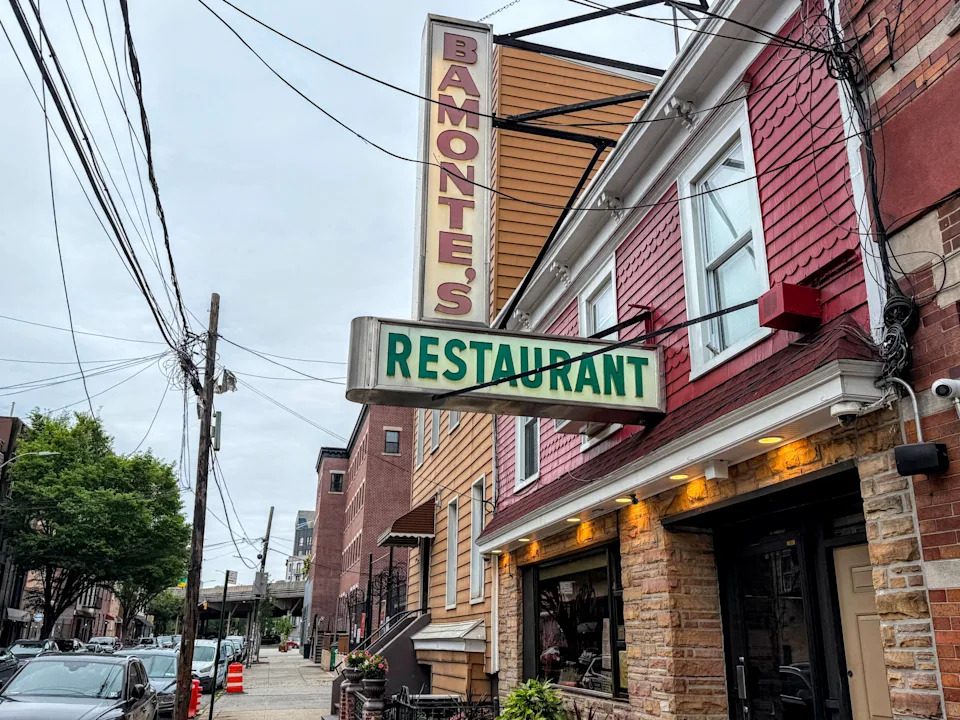 A red-shake building with tan stone facing stands on a narrow Brooklyn street. A vertical white sign reading "Bamonte's" in red letters and a horizontal white sign reading "Restaurant" in green letters are suspended above the entrance.