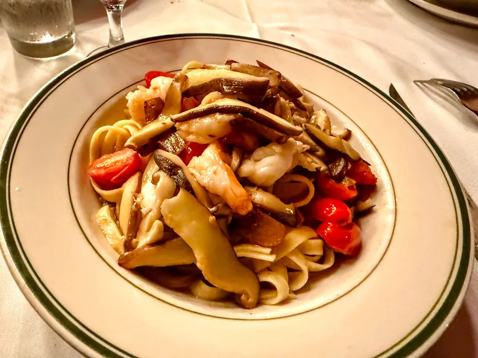 A white dish of fettuccine noodles is topped with sliced, red cherry tomatoes, shrimp, and thinly sliced portobello mushrooms.