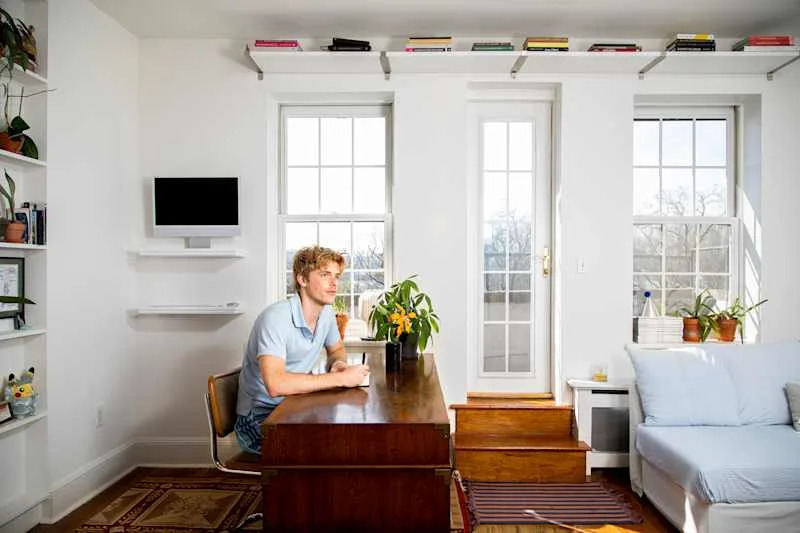 A young man sitting at a wooden desk in a bright room, with plants, a TV, and a staircase leading to a door.