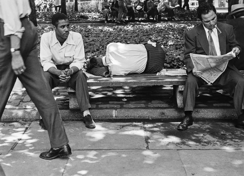 Three figures share a Bryant Park bench: one man sits watchfully, another is slumped face-down asleep, and a suited businessman reads his newspaper with a cigarette in his mouth.