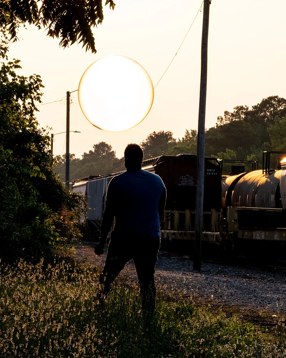 Silhouette of a person standing near train tracks with a bright sun glowing like a large orb in the background at sunset