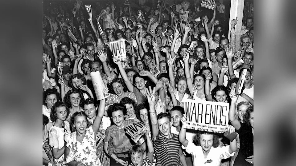 A crowd celebrating. Some people are holding newspapers with the headline