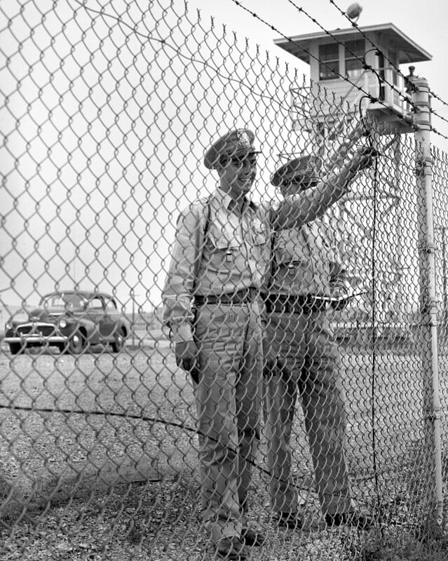 Two uniformed guards stand behind a chain-link fence near a guard tower. One holds the fence; a car and gravel lot are visible in the background. The scene appears to be at a secure facility.