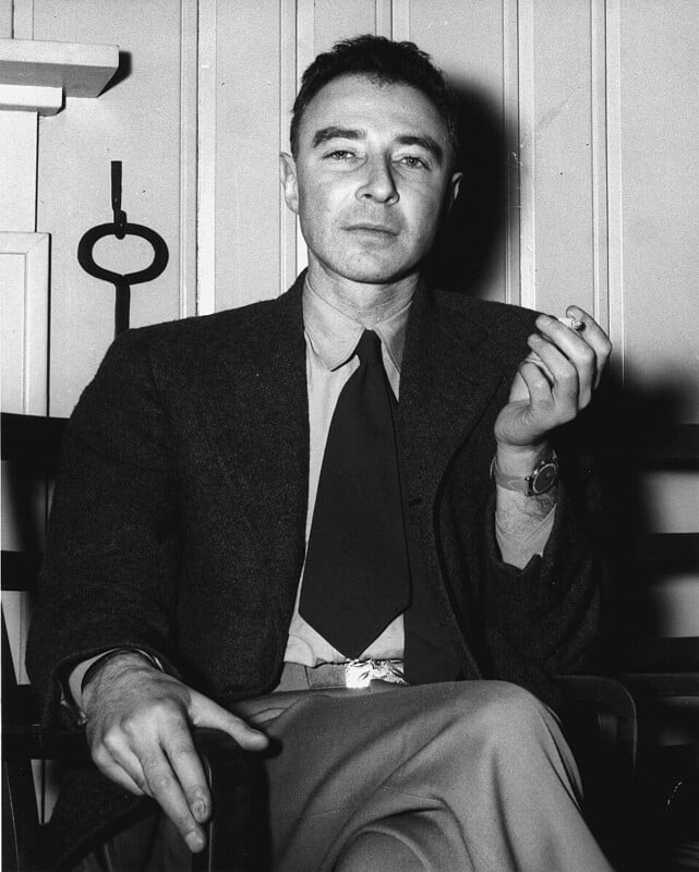 A man in a suit and tie sits in a chair, holding a cigarette in his left hand and looking directly at the camera. He has short dark hair and a calm expression. The background features a paneled wall.