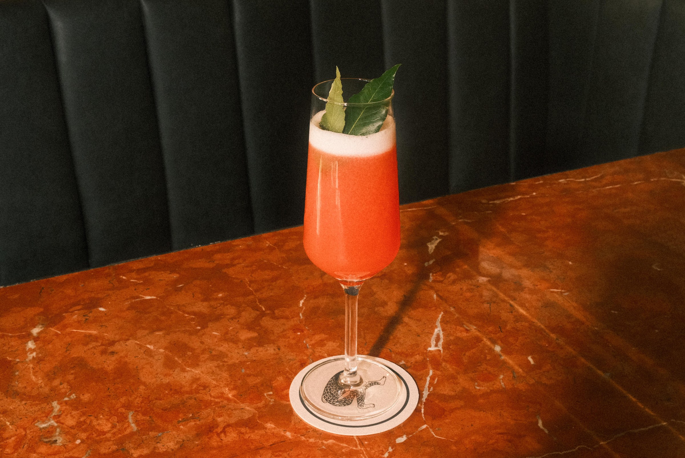 A cocktail with red liquid in a Champagne glass and a leaf garnish.