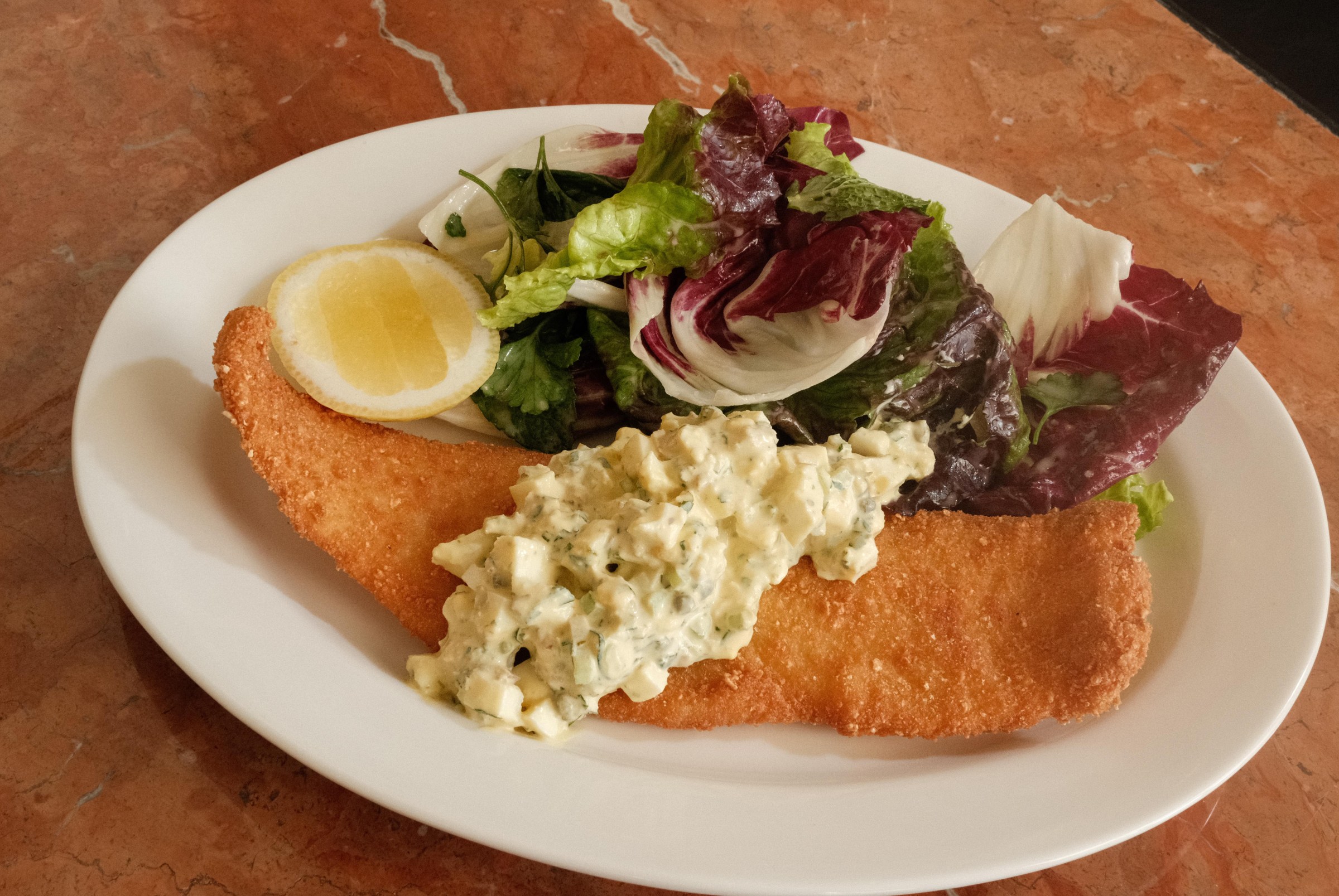 A plate of a long fried fish filet with cream on top of it wit a lemon slice and greens.