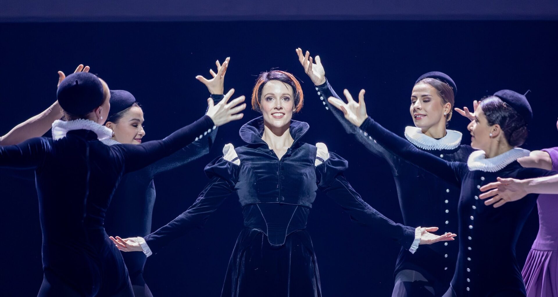 A woman with a short, dark bob with bright red streaks stands and smiles, her arms in demi second with her palms facing up. She wears a black velvet dress with long, puffed sleeves and a high collar. Four women, also in black velvet costumes and hats, surround her with their arms up.