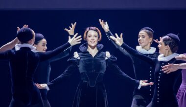 A woman with a short, dark bob with bright red streaks stands and smiles, her arms in demi second with her palms facing up. She wears a black velvet dress with long, puffed sleeves and a high collar. Four women, also in black velvet costumes and hats, surround her with their arms up.