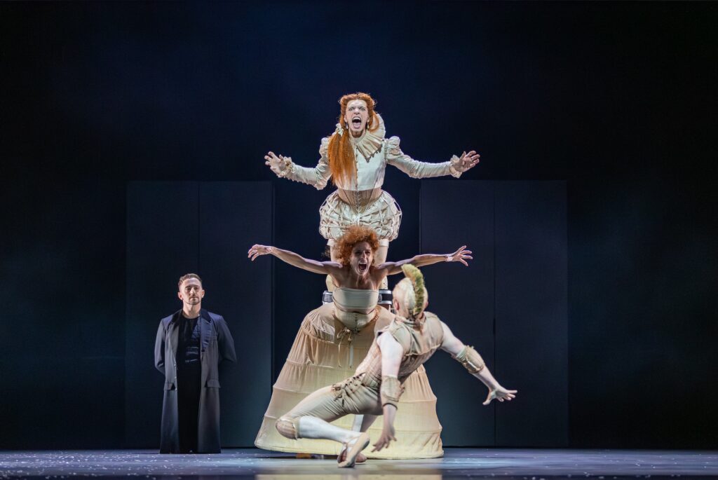 A dancer on stilts stands behind another dancer. They both wear Elizabethan costumes and reed wigs, and scream with their arms out to the side. Another dancer faces them and falls backwards in fear.